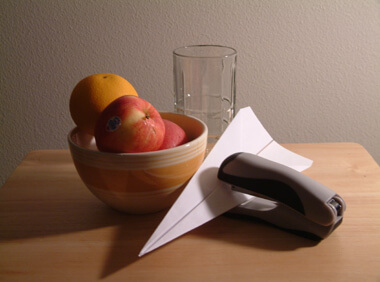 Still life setup of bowl with fruit, glass, paper airplane, and stapler. The strong lighting from the side makes it easier to draw.