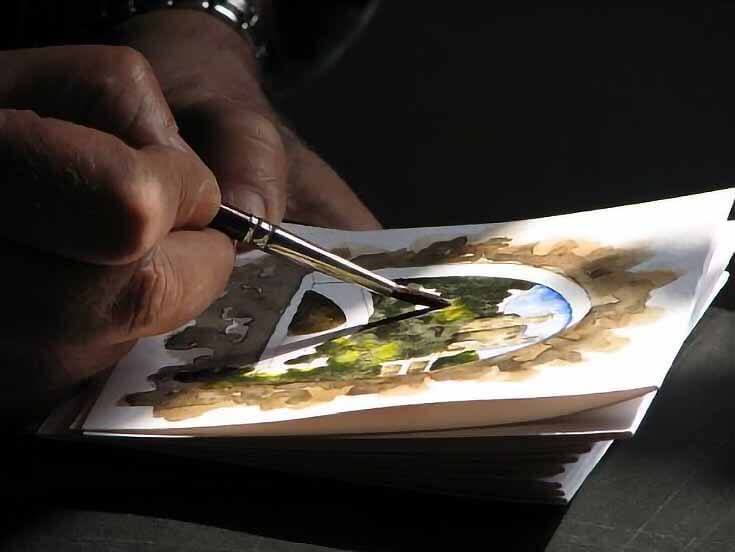 Close-up photo of a hand holding an artist's brush, painting a small watercolor scene with trees and buildings.