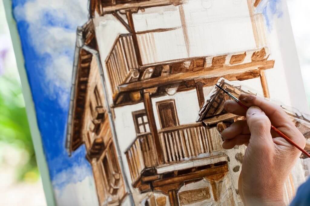 Photo of a hand holding a brush, making deliberate marks on a painting of a house