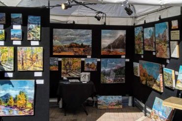 Artist booth in outdoor art show with paintings hung on a black background